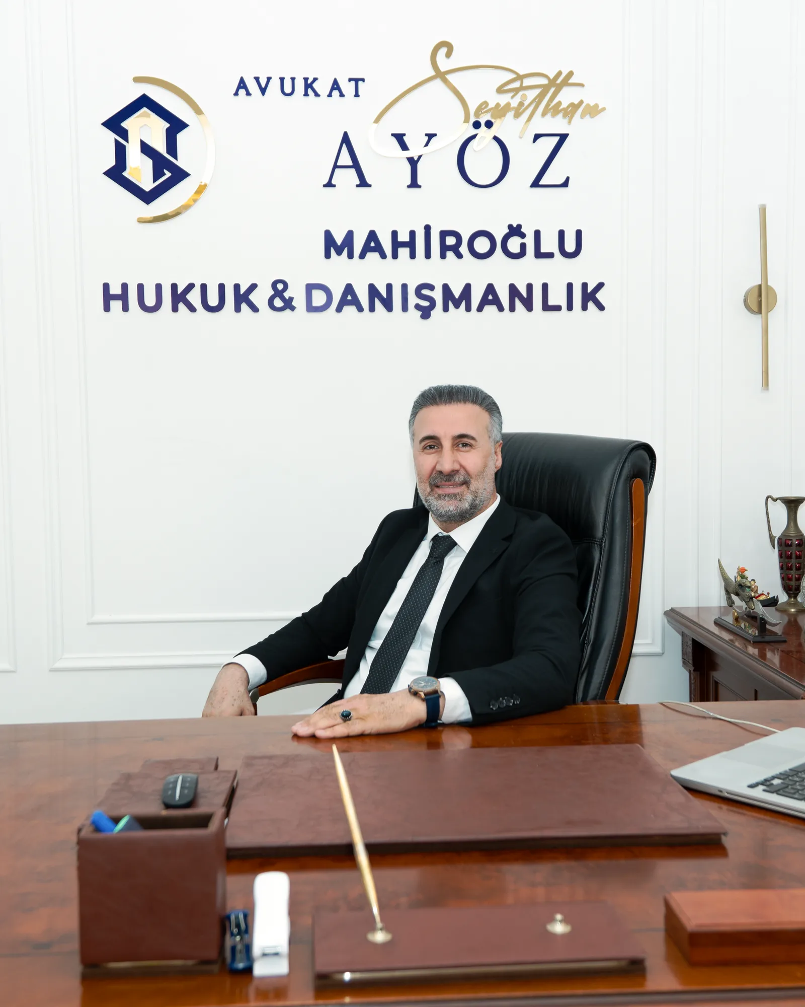 Mahiroğlu Hukuk & Danışmanlık avukatı, büro logosunun önünde makam masasında