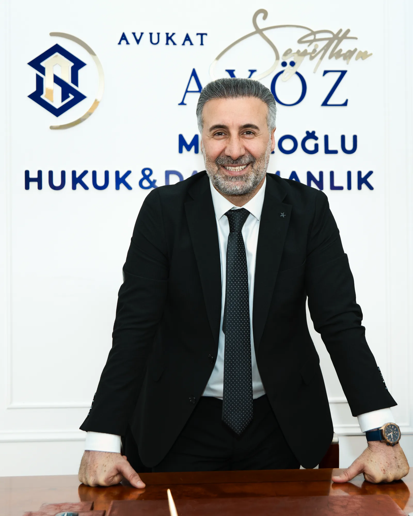 Mahiroğlu Hukuk & Danışmanlık avukatı masaya yaslanmış, gülümseyen ayakta portre
